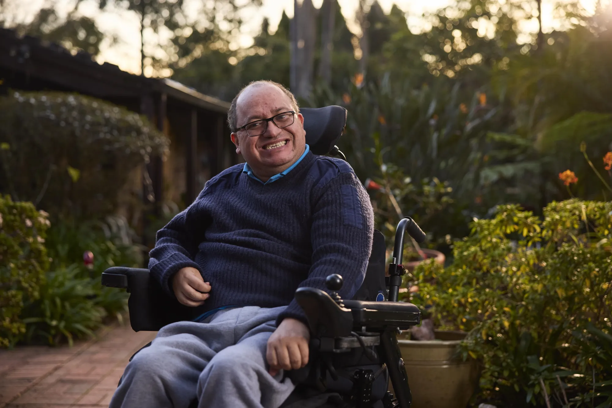 A person in a wheelchair sits outdoors amidst lush greenery, wearing a dark sweater and grey pants, enjoying the warm evening light.