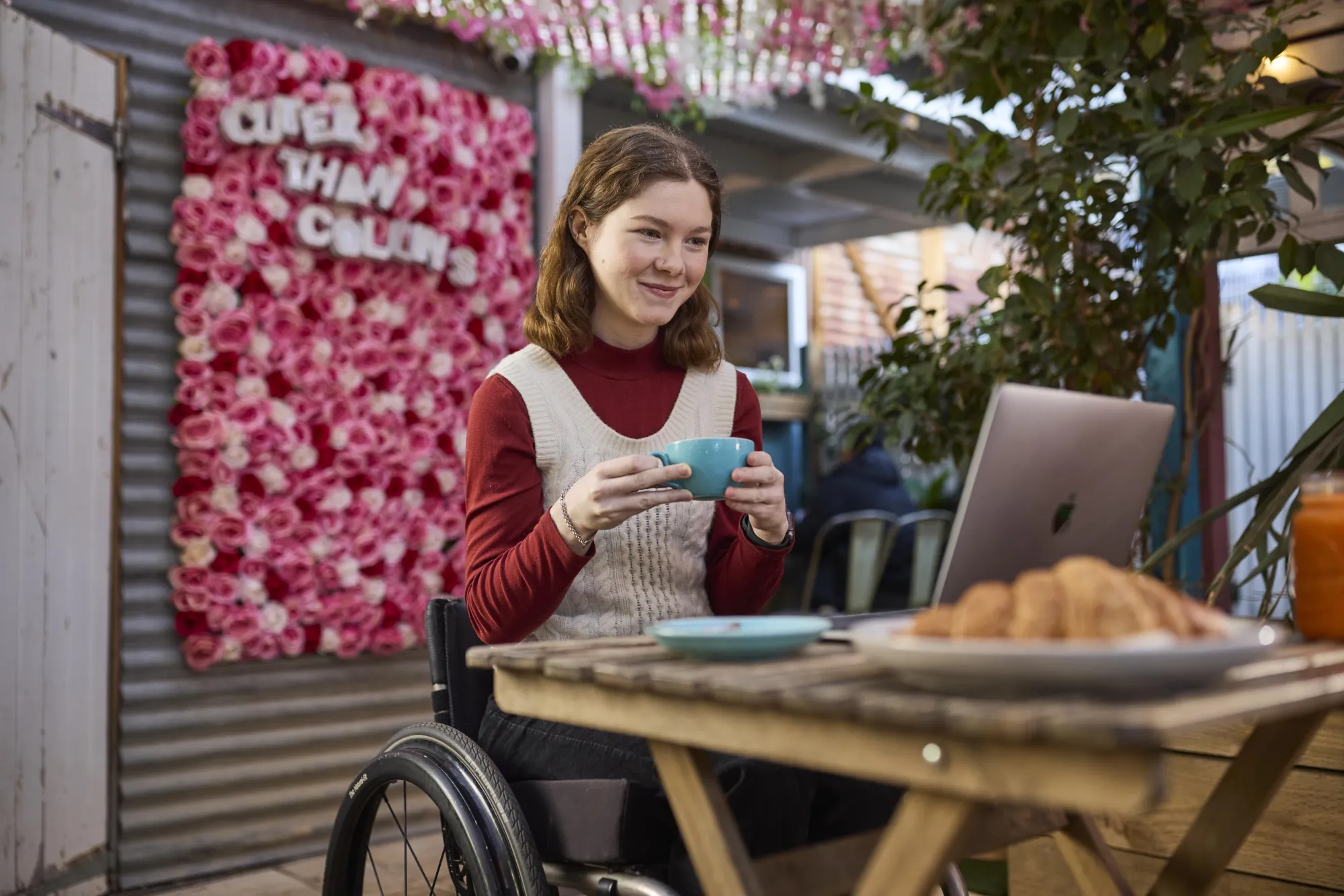 A person in a wheelchair holds a blue cup, sitting at a wooden table with croissants, against a backdrop of pink floral decorations.