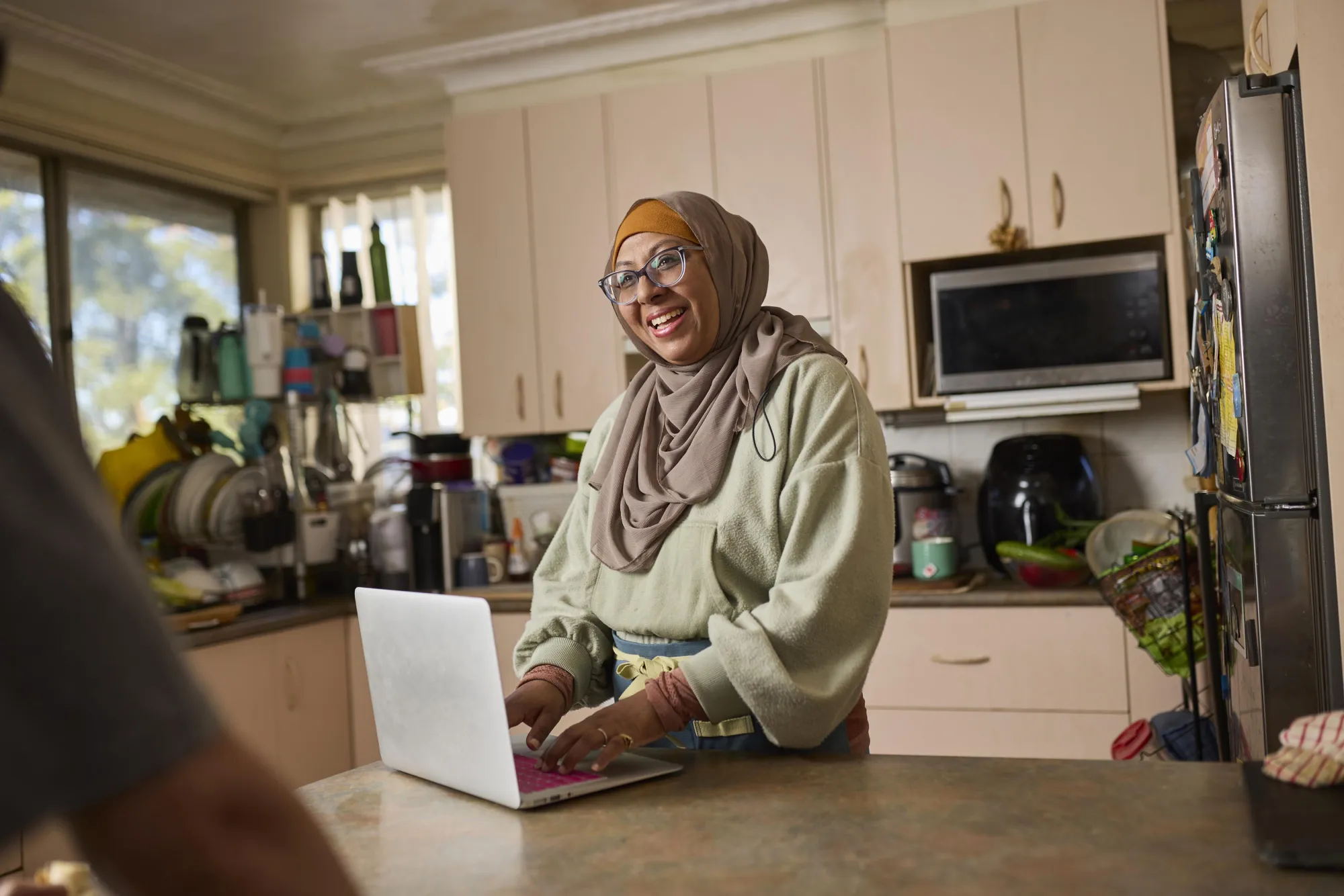 A person in a cozy kitchen, wearing a light green sweater and beige head scarf, types on a pink laptop near various kitchen items.