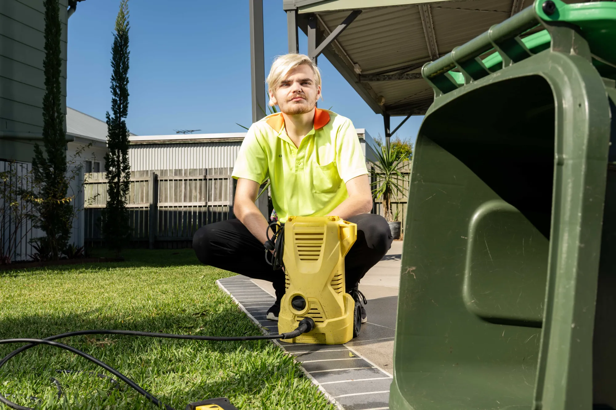 A person who works a physical job in an outdoor setting. They have a yellow high vis shirt on. There is a bin in the foreground of the photo. The bin is Forrest green. 
