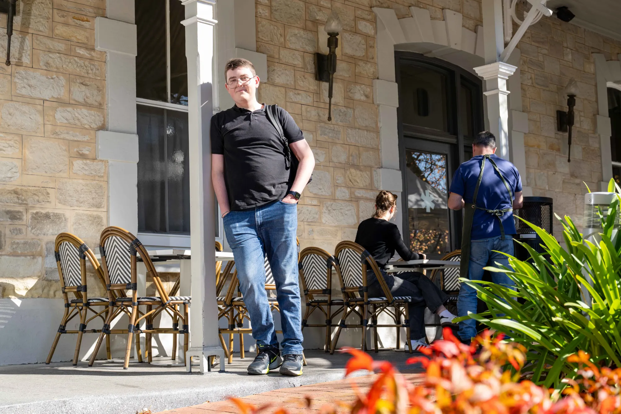 A young man standing against a café, with his hands in his pockets. They are smiling. 