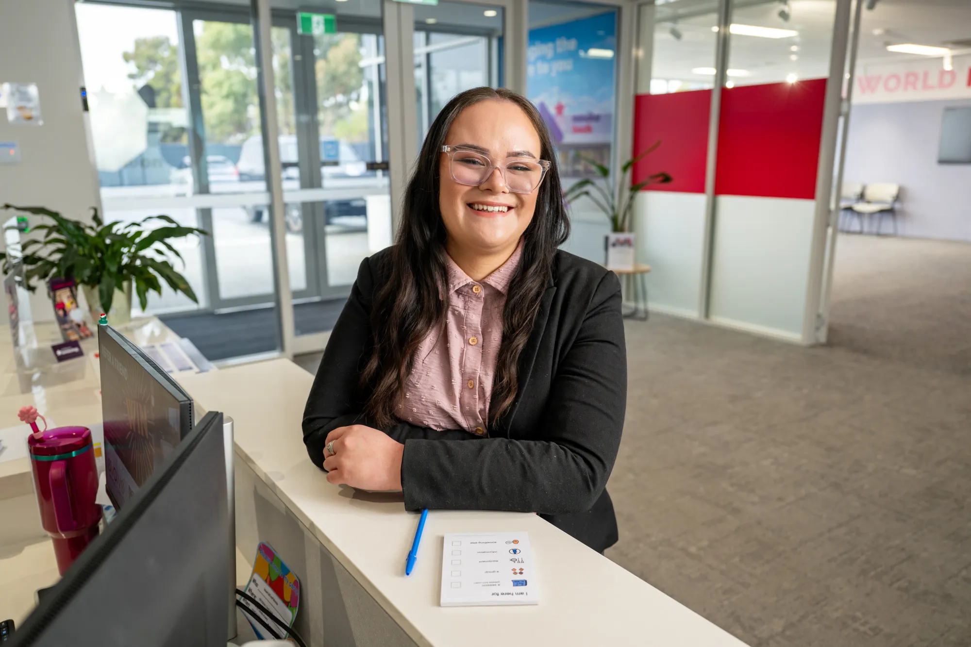 A young worker leaning on the front desk of a office. They have clear glasses and a smile on their face. 