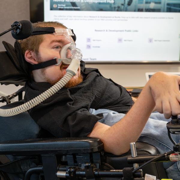 A person in an electric wheelchair with a face mask to assist with breathing. The person has a smile while being in an office setting.