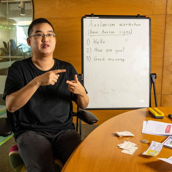 A person using Auslan sign to explain something. Behind them is whiteboard with some basic of Auslan.