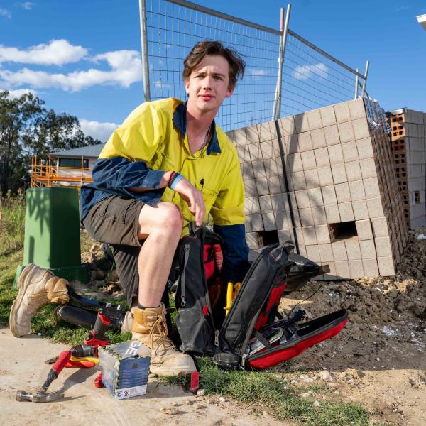 A young tradesperson with a kneeling down on a construction site. They have a prosthetic on their left foot. 