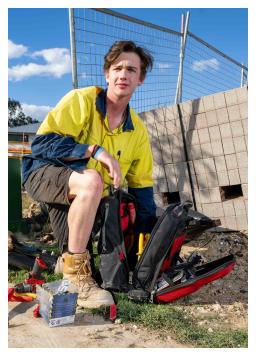 A young tradesperson with a kneeling down on a construction site. They have a prosthetic on their left foot. 